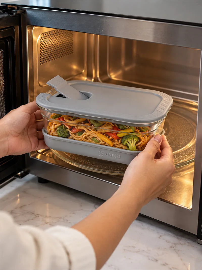 A person's hands holding a clear glasafe  glass meal prep container with a gray lid and a small white flip-up tab, containing noodles and broccoli, as it is placed inside a stainless steel microwave oven.