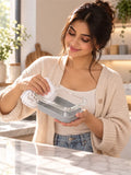 A woman in a beige cardigan and white tank top holds a clear glass rectangular container with a gray silicone liner while wiping it with a white cloth in a bright kitchen setting.